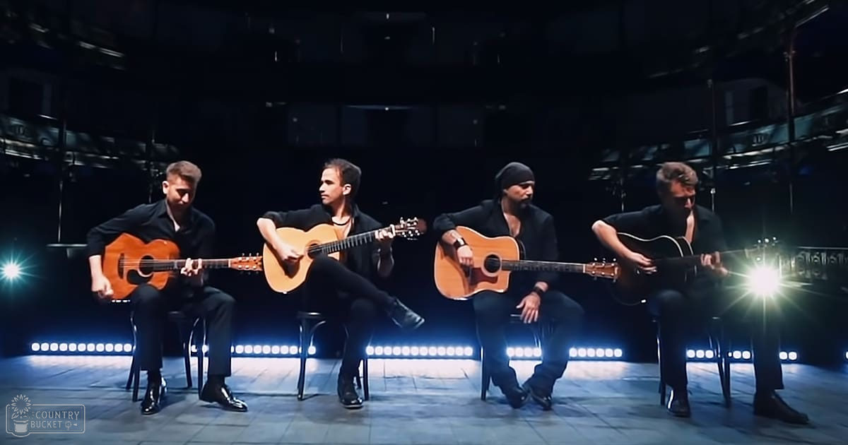 Musical performance scene with multiple adult males in dark blazers, seated on black chairs under bright stage lights and industrial-style ceilings at night.