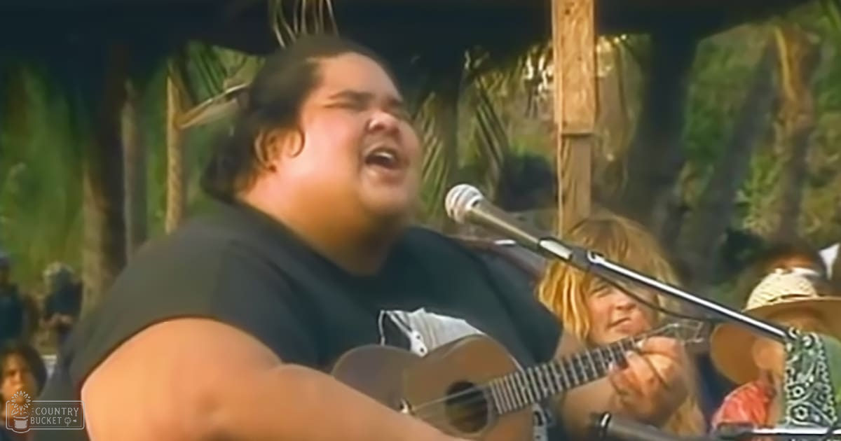 A young adult male singer wearing a black sleeveless shirt plays guitar while singing into a microphone at an outdoor festival with palm trees and wooden structures, suggesting daytime summer weather.