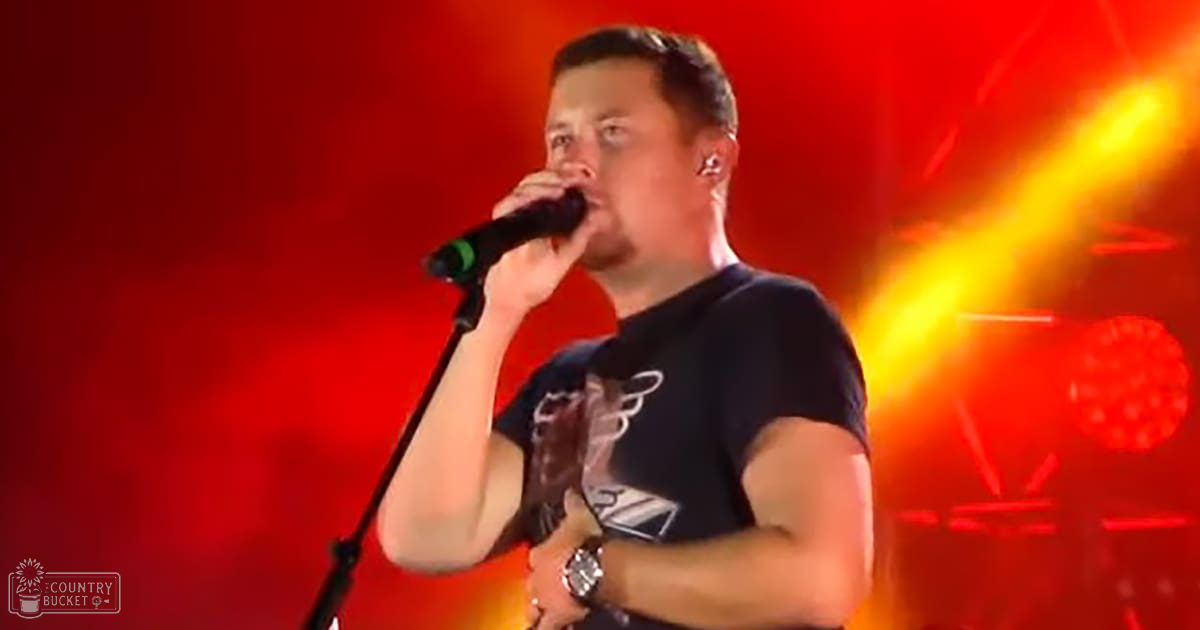 An adult male singer wearing a dark t-shirt with a graphic design, holding a microphone and wristwatch, performing at an indoor concert venue with dramatic stage lighting.