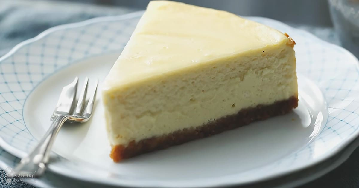 A slice of cheesecake with a fork on a plate, set against a backdrop of a blue and white checkered cloth in an indoor kitchen setting