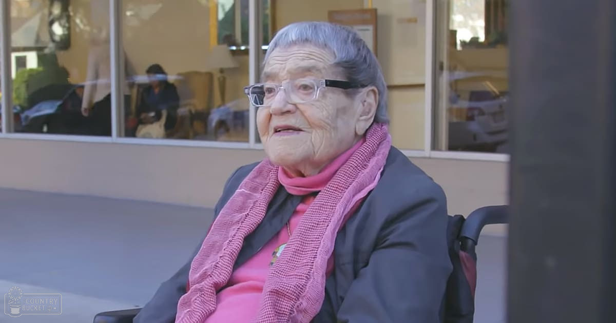 An older adult female in a wheelchair outside a building, wearing a pink sweater and glasses with a dark jacket layered under it.