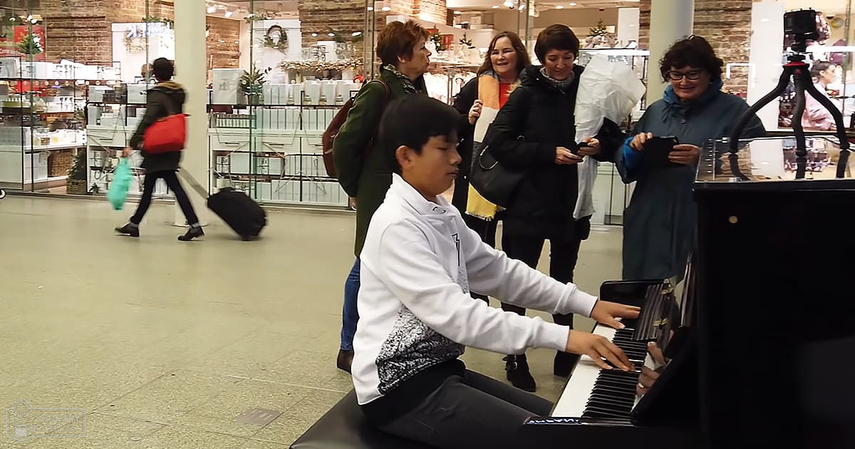 Young adult male playing piano in an indoor shopping area with others watching; winter clothing styles, natural light.