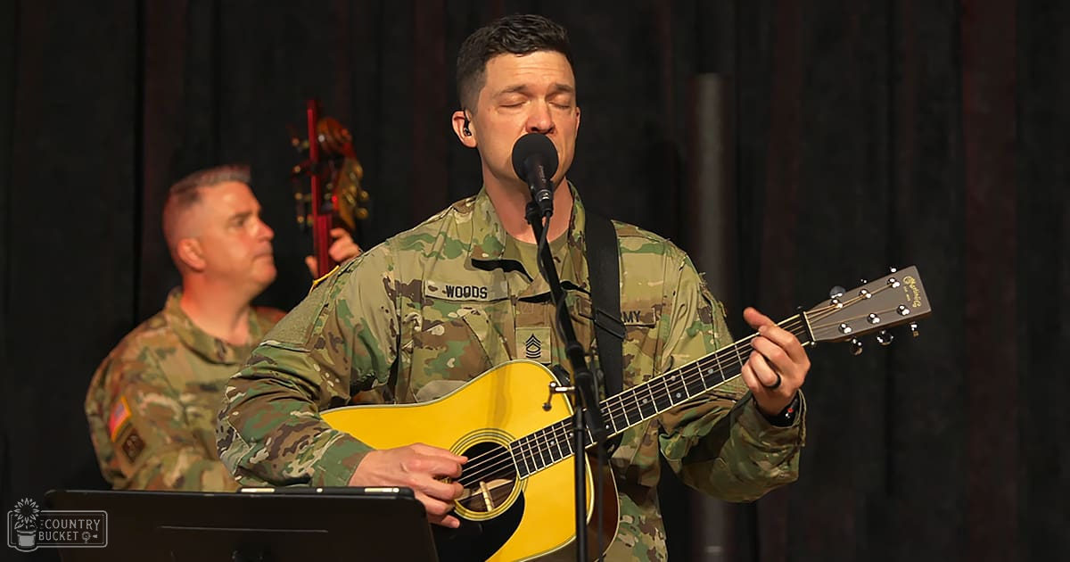 A soldier performing music on stage with another military member in the background under artificial lighting at night.