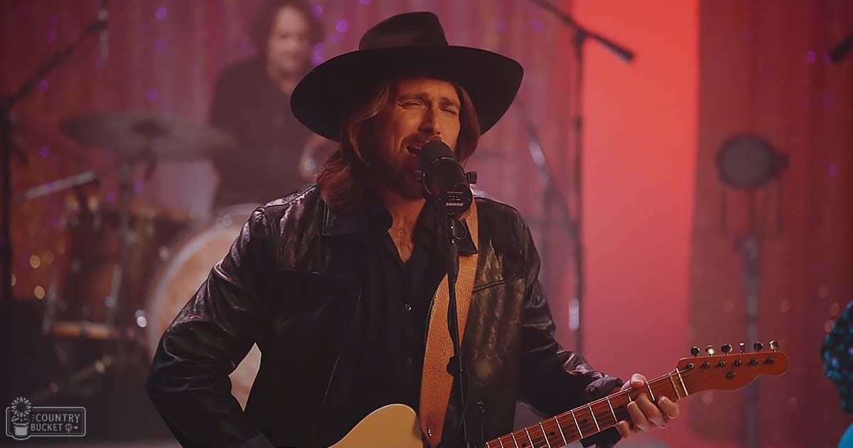An adult male musician wearing a black leather jacket with high collar and zip, performing on stage at an indoor concert venue with red curtains and purple lights in the background.