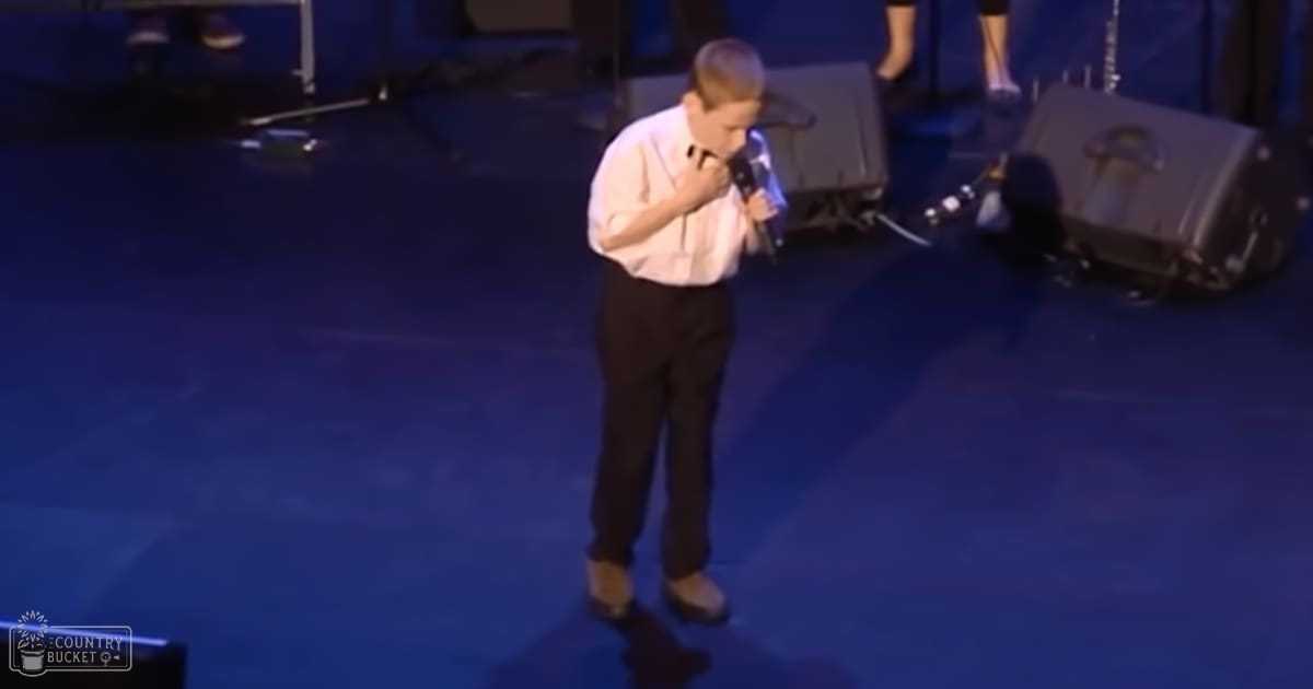 A young adult male in a white button-up shirt, black trousers, and brown shoes stands on stage with a microphone, surrounded by equipment and part of another individual's legs. The setting suggests an indoor event or performance.