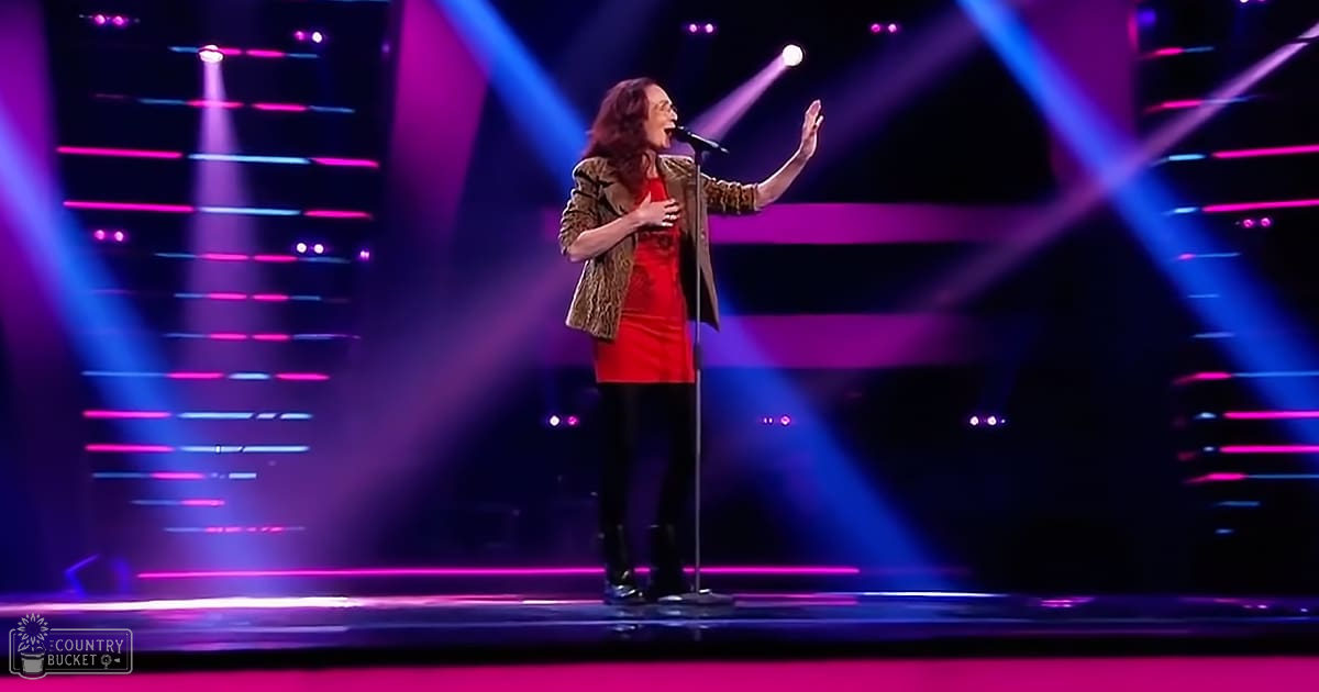A middle-aged female performer in a red, fitted dress with long sleeves, singing or speaking on stage under vibrant pink and blue lighting.