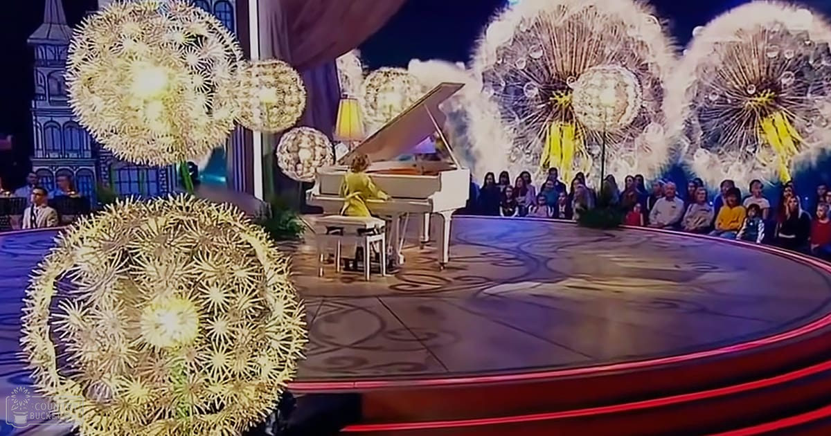A grand piano on a stage with an audience in various attire, indoors at what appears to be a concert or performance venue.