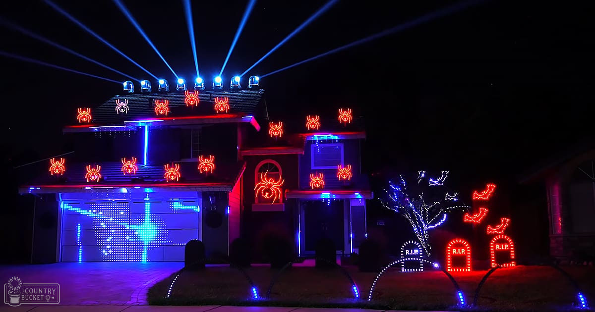 Halloween decorations, illuminated spider figures, tombstones 'R.I.P.', blue laser lights at night in suburban neighborhood.