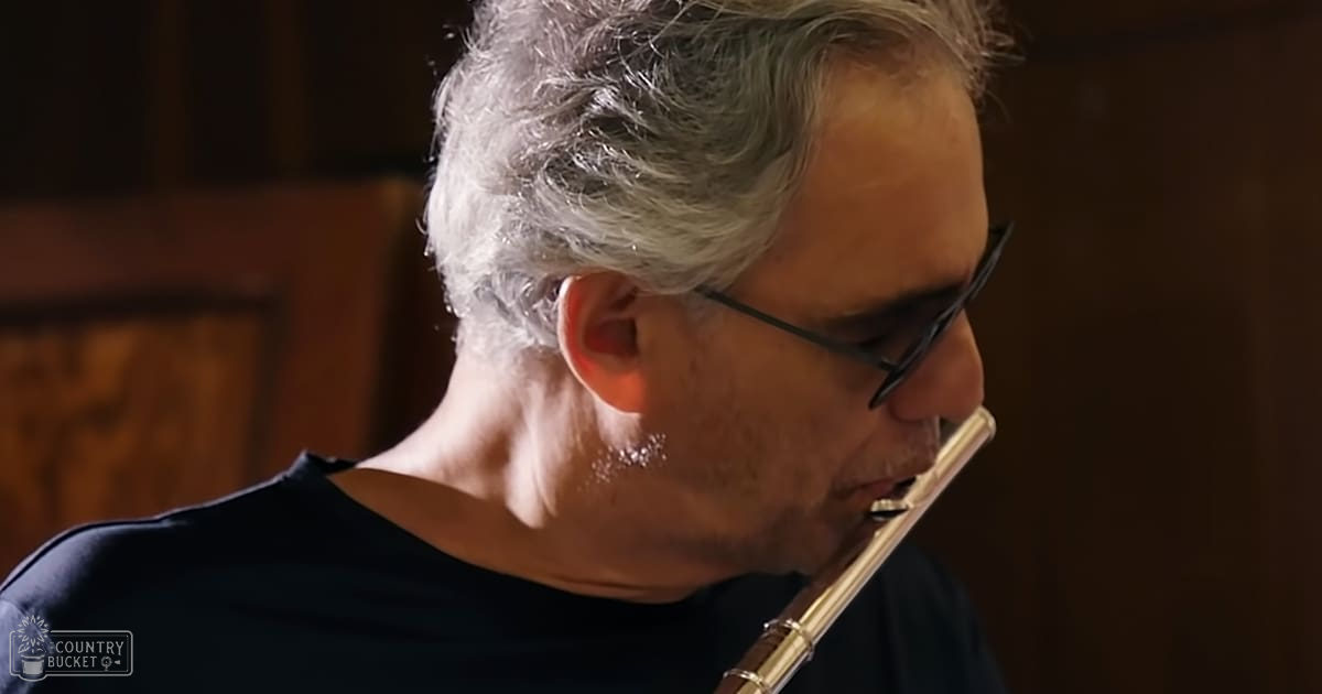 A middle-aged adult male playing a flute in an indoor studio with warm-toned wooden panels and soft lighting.