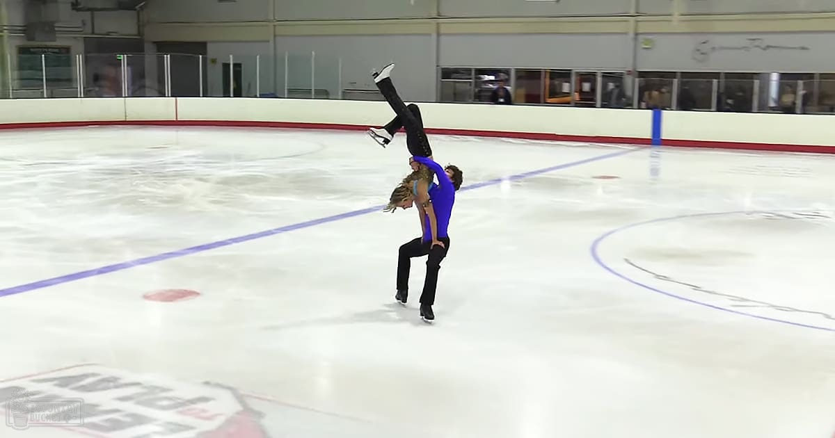 A young adult female figure skater in a blue leotard performs alongside an adult male figure skater at an indoor ice rink with markings and signage visible in the background.