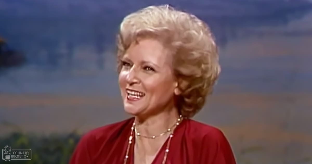 An older adult female wearing a red, loose-fitting, low-cut neckline, bell-sleeved garment with pearl jewelry, set against an even-lit studio backdrop.