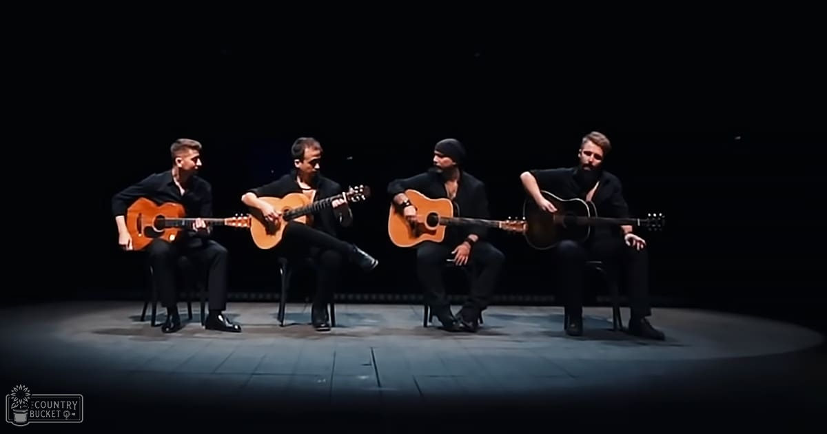 A musical performance with four adult male musicians seated and playing acoustic guitars in an indoor stage setting at night.