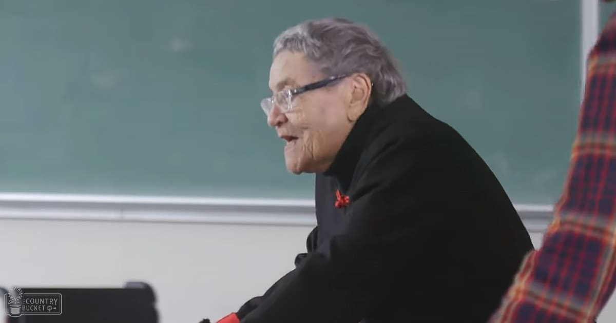 An older adult female in a classroom setting with short gray hair wearing glasses and a dark garment, surrounded by educational elements.