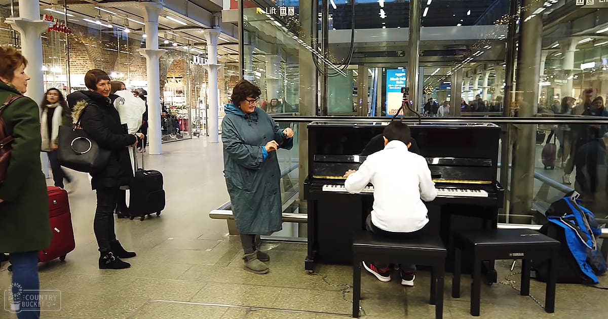 A young boy plays the piano in an indoor public space resembling a train station or shopping mall, surrounded by adults wearing winter clothing.
