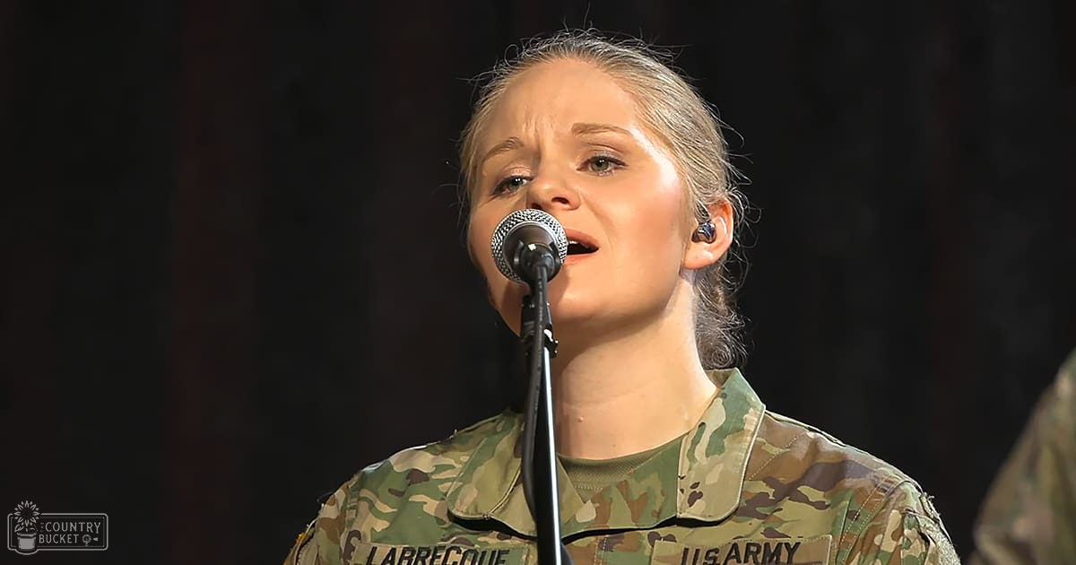 A young adult female in a U.S. Army camouflage uniform sings into a microphone on an indoor stage under dim lighting.