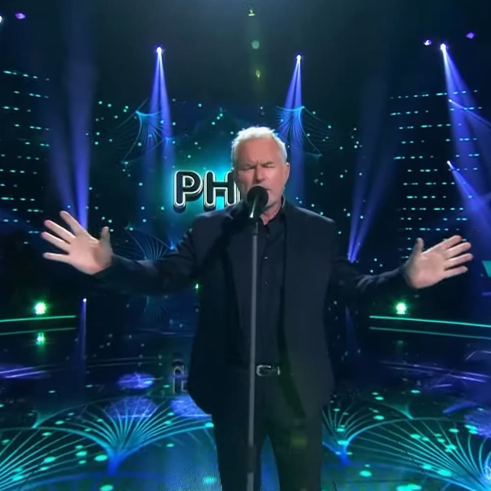Middle-aged male performer in a dark suit at an indoor stage with professional lighting, wearing no visible footwear or accessories. A microphone stand and decorative blue and green lights are present behind him.