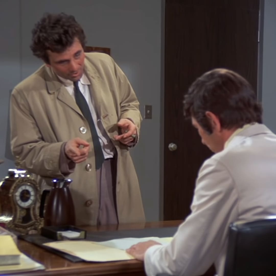 An adult male with curly hair, wearing a beige trench coat over a white shirt and dark tie, standing indoors in an office setting.