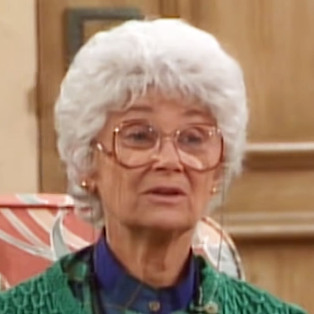 An older adult female wearing a green, textured sweater with long sleeves and glasses in a domestic setting with wooden furniture and natural light.