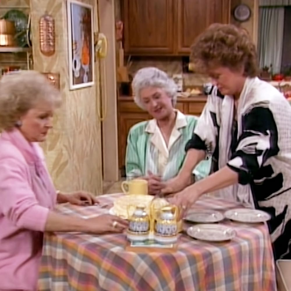 Three older adult females in a cozy kitchen setting from 'The Golden Girls,' interacting around a wooden table with teapots and plates.