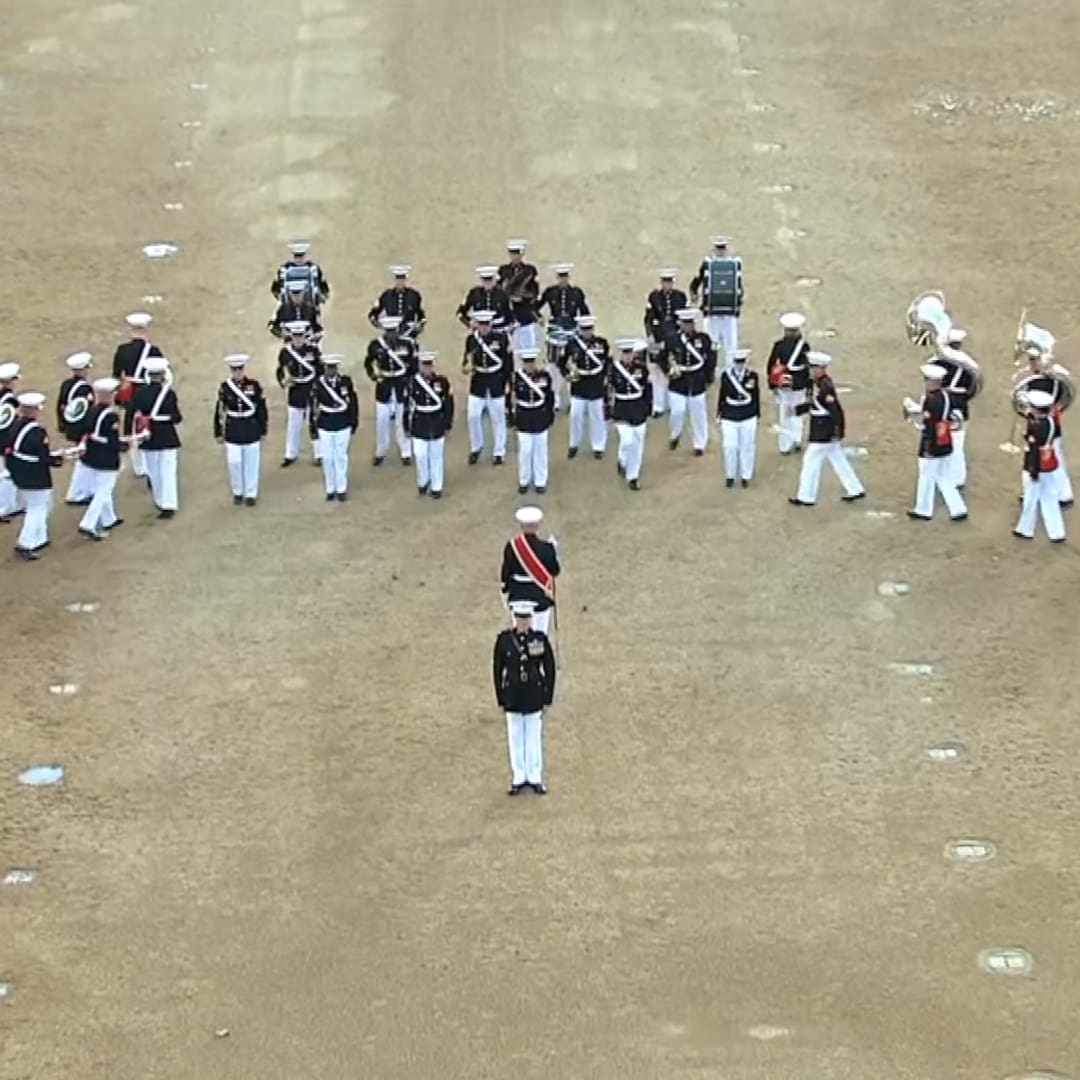 A military parade on a sandy field under bright daylight, with young adult male soldiers in dark uniforms and white trousers, some wearing red sashes indicating rank.