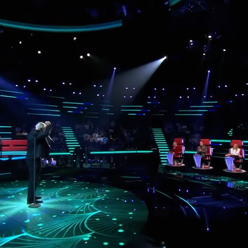 A male older adult singer wearing a dark suit jacket, white patterned tie, and black shoes, holding a microphone on an indoor talent show stage with judges seated in red chairs at night.