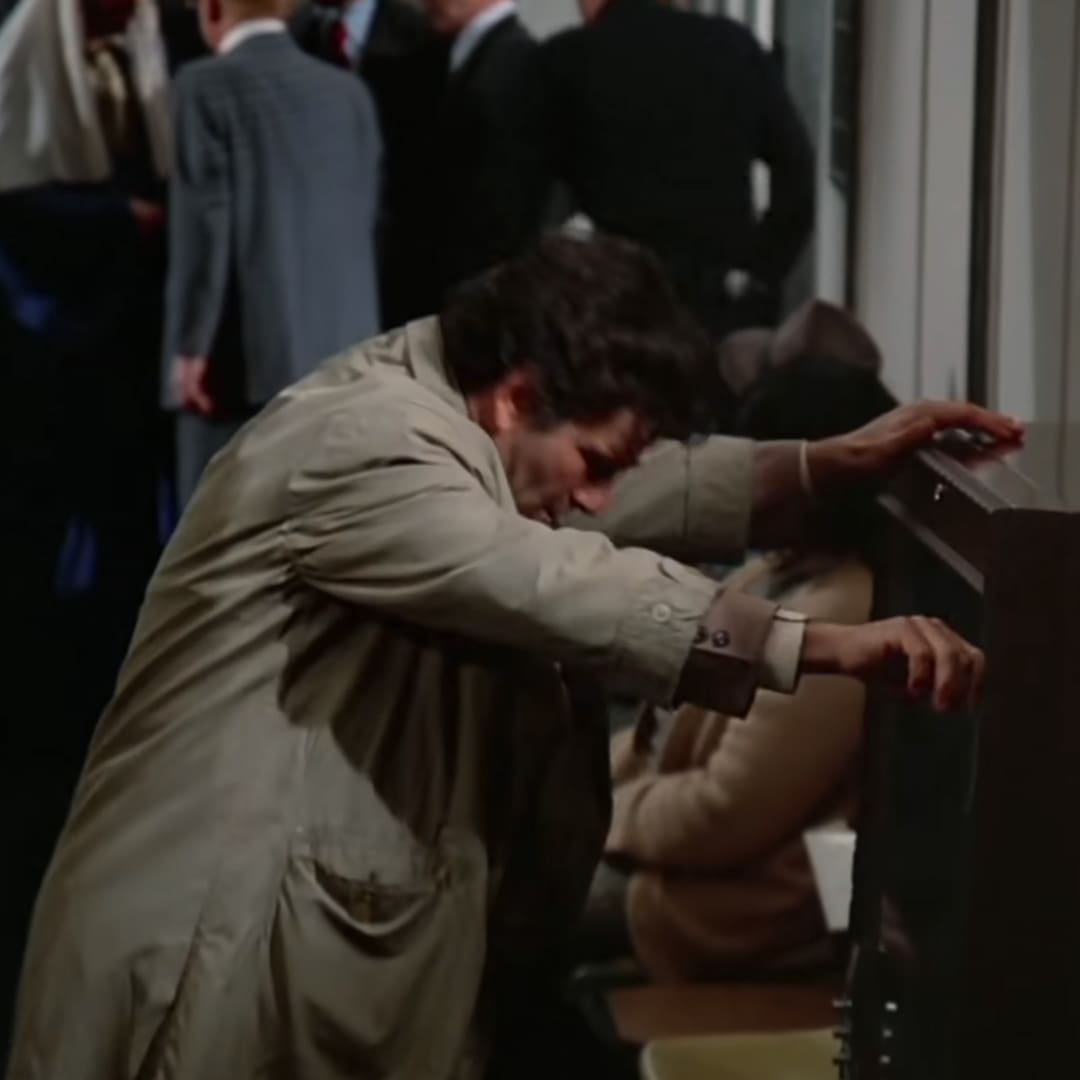 A man in a beige trench coat with large pockets, possibly mid-20th century vintage style, stands indoors near dark furniture. Other adults in business attire are present, suggesting an office or formal meeting room setting.