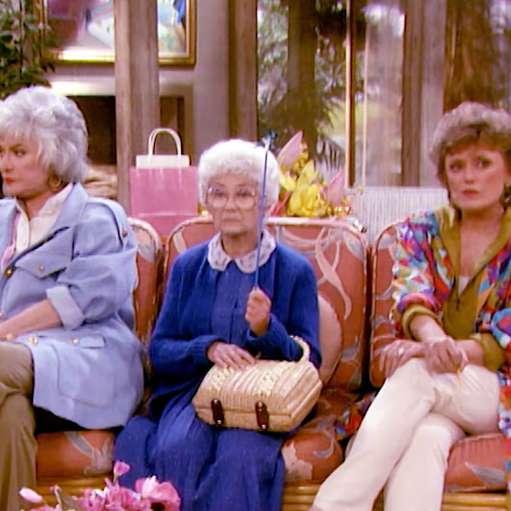Three adult female characters seated indoors, wearing trench coat, sweater with white shirt and dark pants, floral blouse over vest; holding umbrella, cross-legged on patterned sofa with orange and cream tones. Pink shopping bag, flowers, framed picture in the background suggest a cozy living room setting.