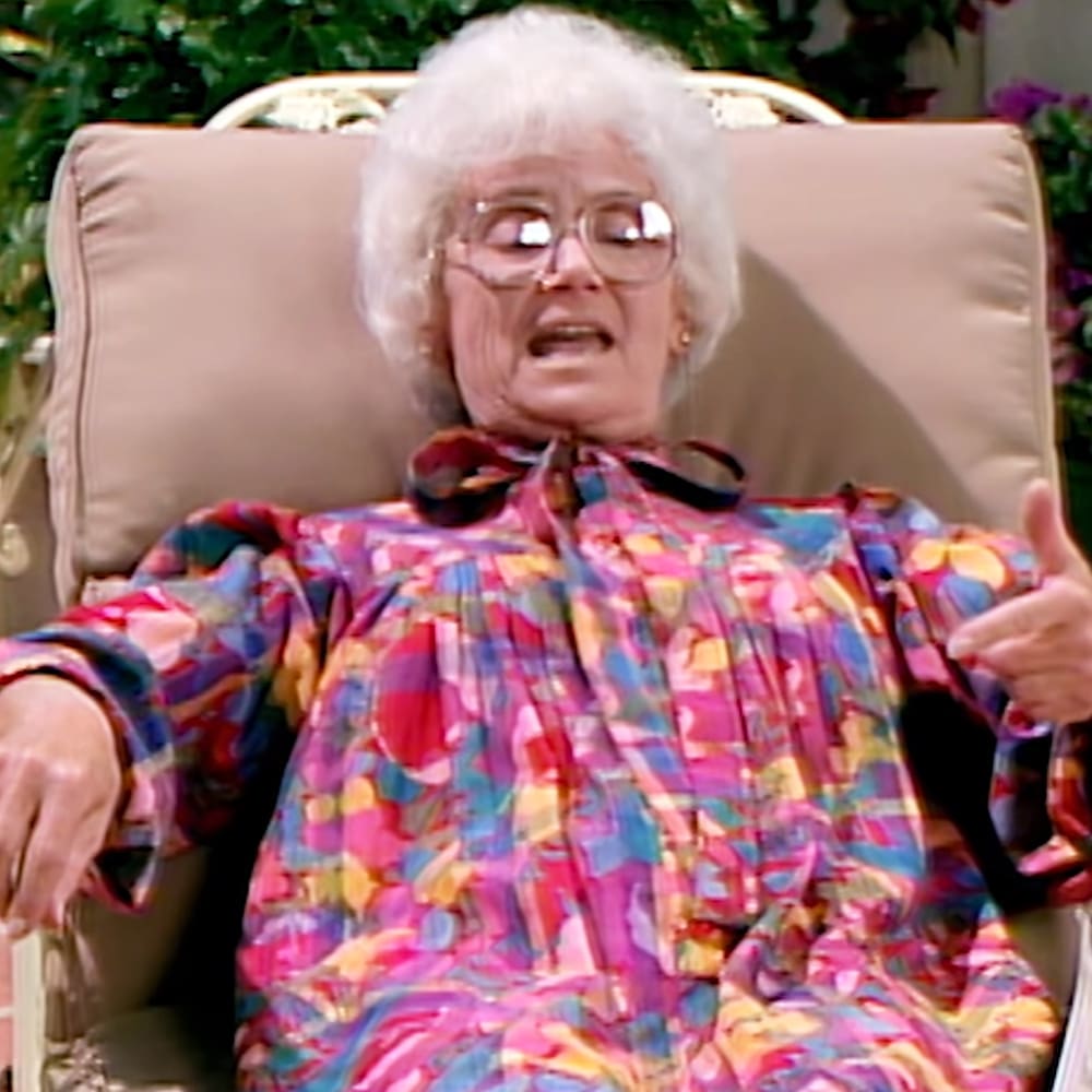 An older adult female wearing a loose, ruffled, bright pink, blue, yellow, and white patterned shirt or dress indoors with beige cushioned furniture and green foliage in the background suggesting daytime light.