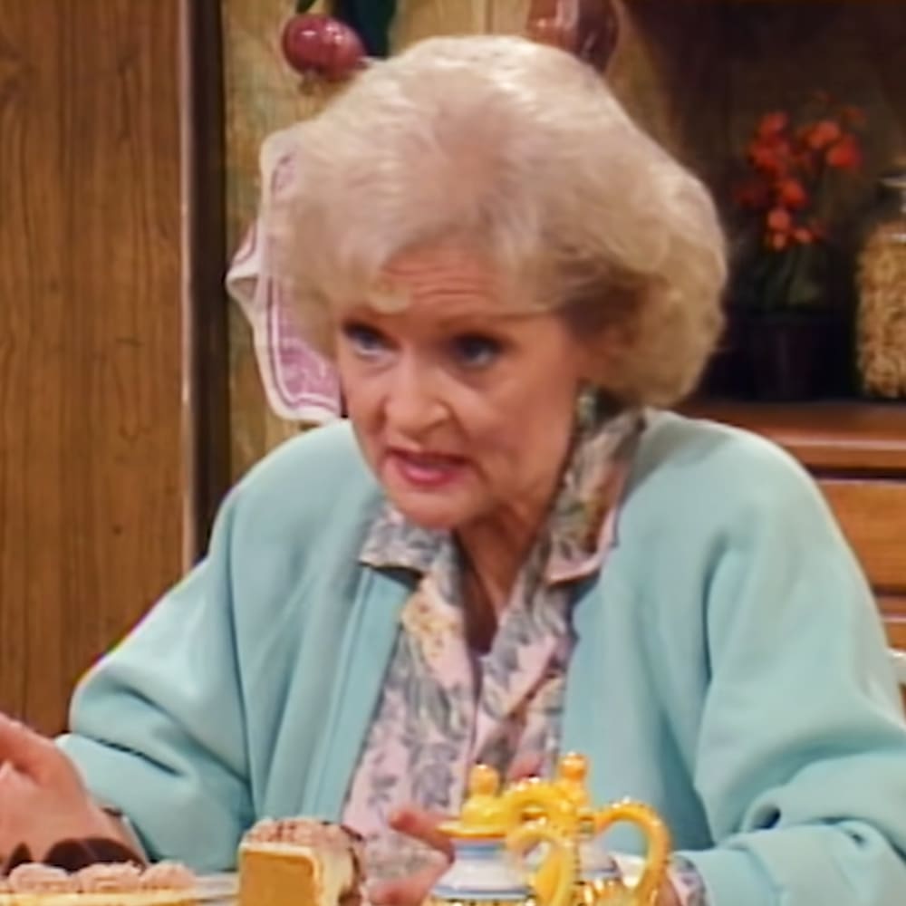 An older adult woman wearing a light blue cardigan with a loose fit, standing indoors amidst kitchenware and home decor items.