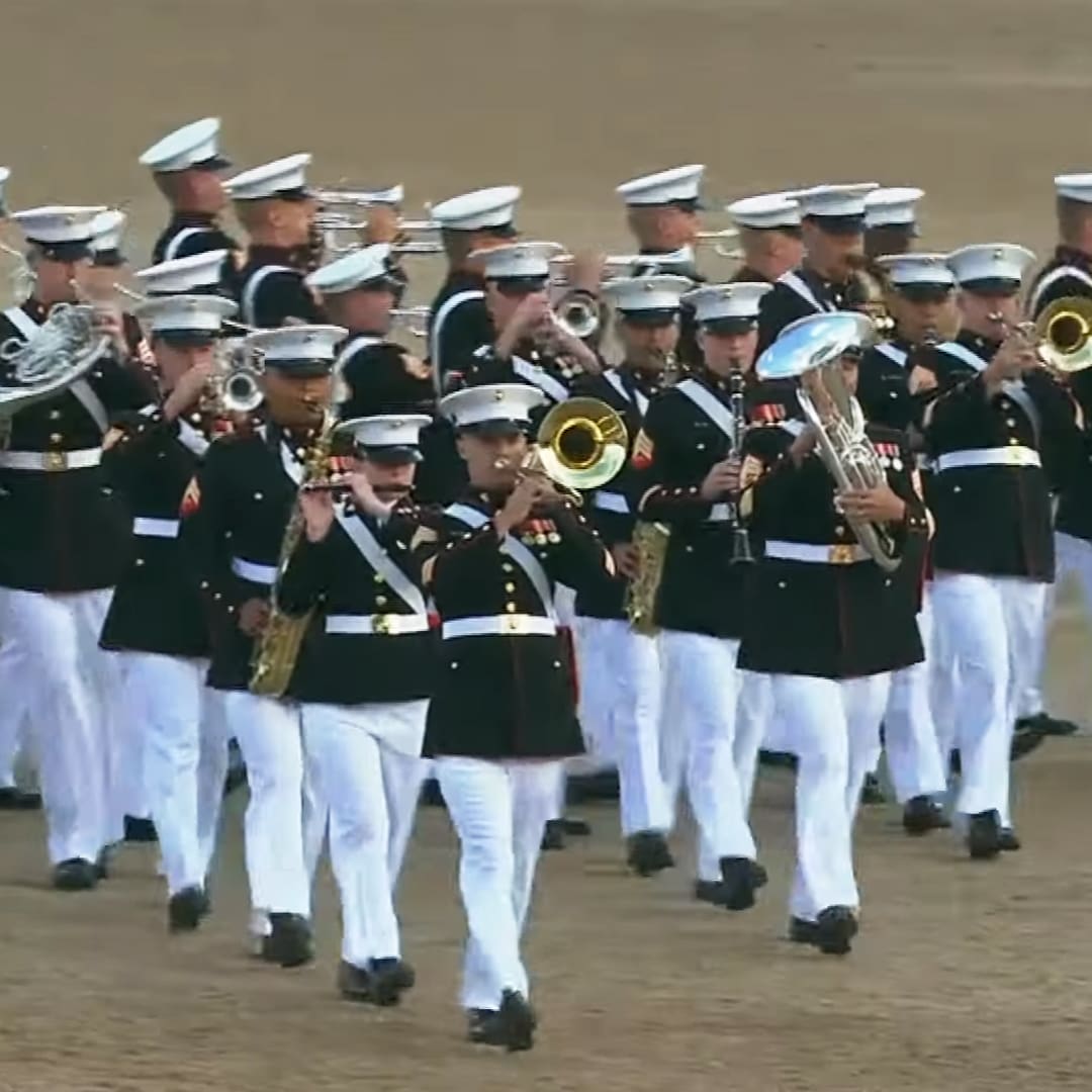 A marching band in military-style uniforms at an outdoor event with clear skies and a dirt ground.