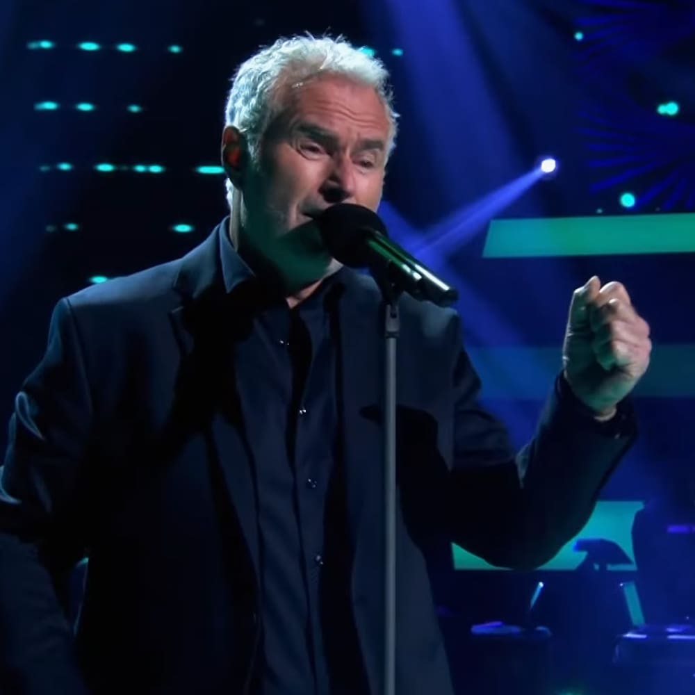 A middle-aged male singer in a dark suit jacket performing on stage under blue and white lighting, likely during an evening concert.