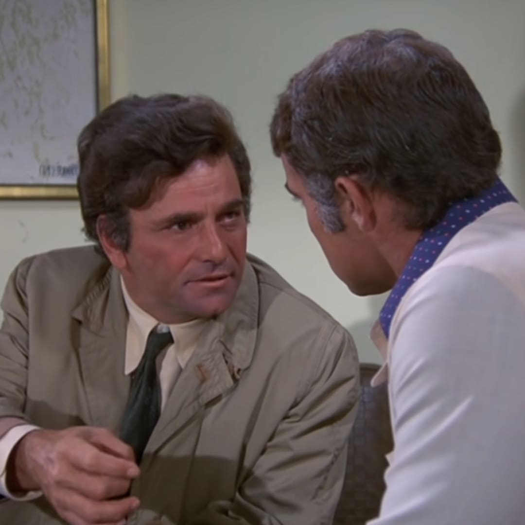 Middle-aged men in a professional indoor setting, discussing or presenting, with light brown blazers and blue-patterned ties against neutral-colored walls.