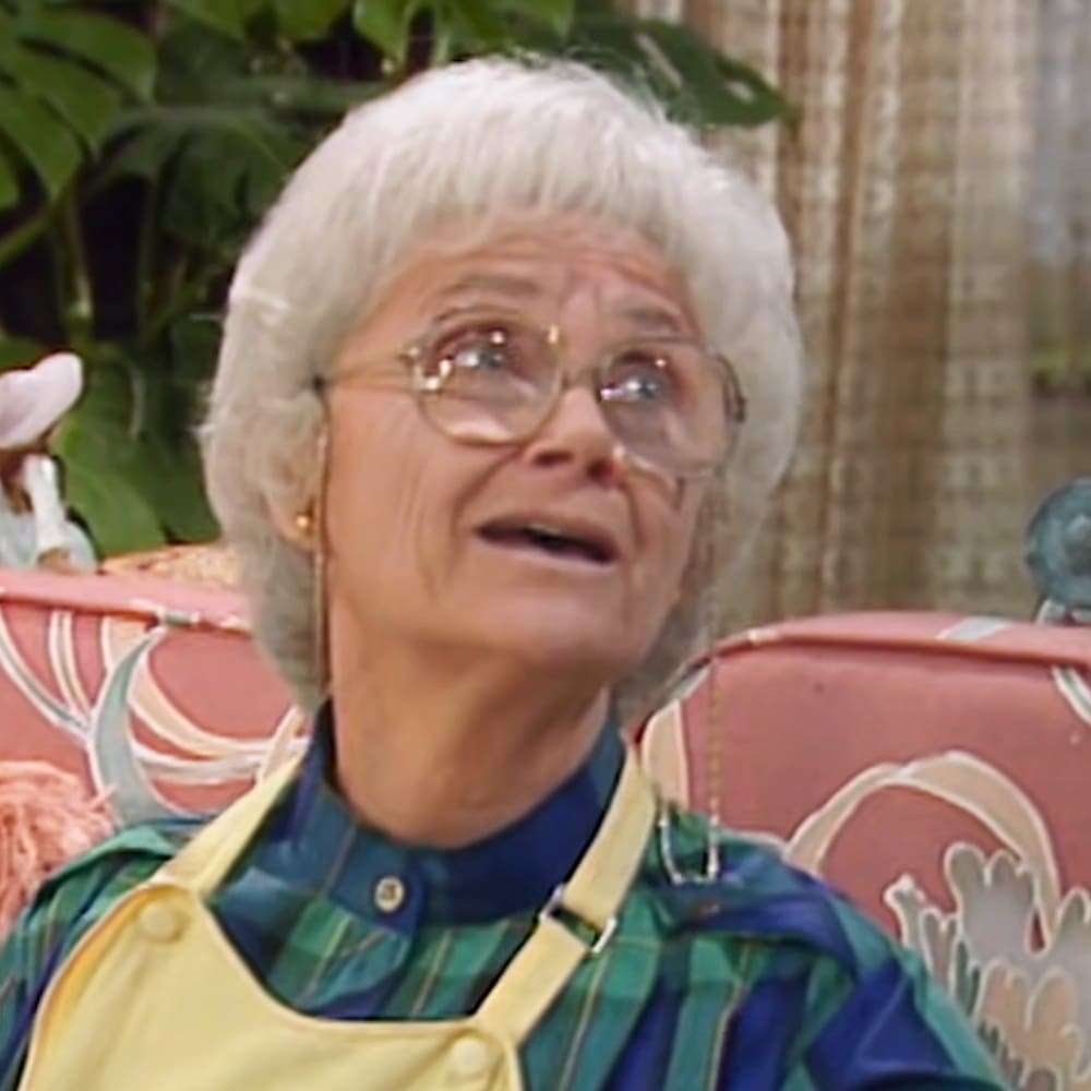 An older adult female wearing a green and blue plaid shirt layered under a yellow apron, seated indoors with a red floral-patterned chair in the background.