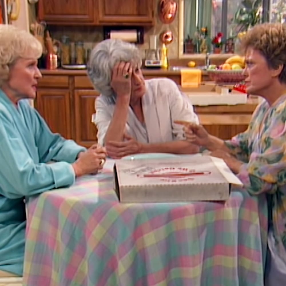 Three older adult females gathered around a kitchen island in a casual setting, wearing various garments and interacting with each other.