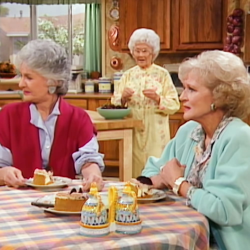 An older adult female character wearing a red cardigan and light blue shirt sits at a wooden table covered with a plaid tablecloth in a kitchen setting, surrounded by another woman in a yellow floral dress and a third woman in a light blue jacket. The scene is bright and natural, suggesting daytime during spring or summer.