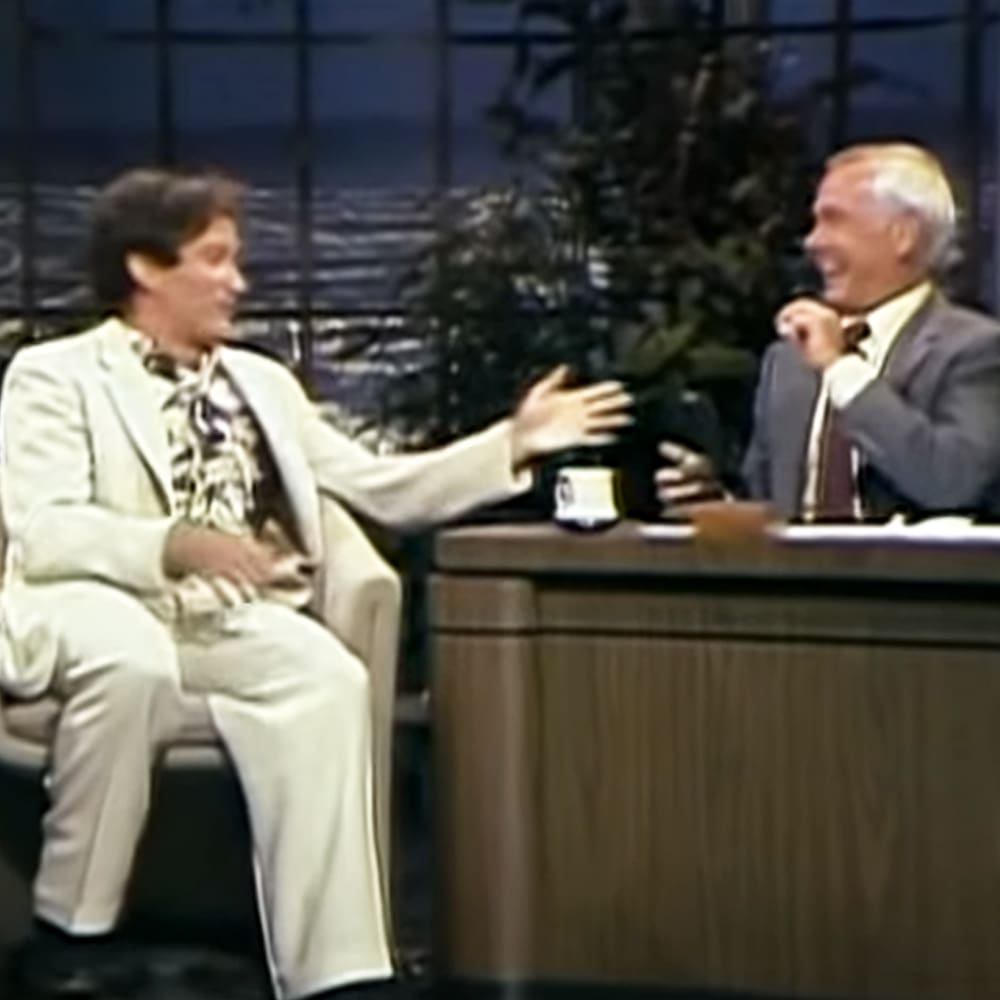 A talk show host and a guest dressed in suits, seated at a desk with decorative items, in an indoor studio setting with dark curtains and greenery.