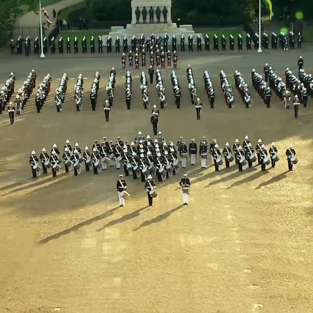A military parade with young adult to middle-aged male soldiers in dark green uniforms and white accents, standing under a monument with statues of soldiers on an open area during daylight likely spring or summer.
