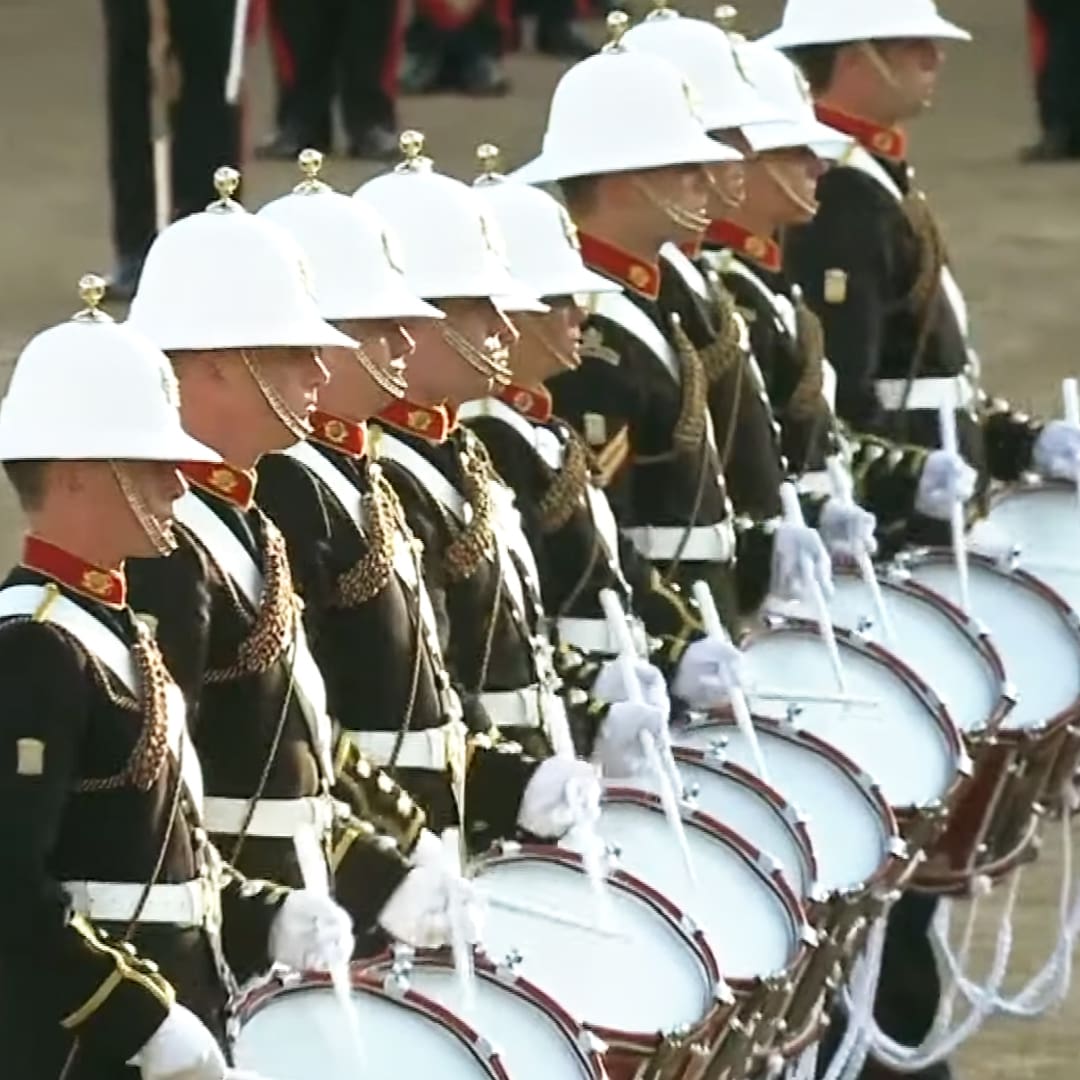 A military parade in an urban setting with young adult male soldiers wearing dark uniforms and white helmets. Each soldier holds a bass drum with white drumheads, red trim, and white drumsticks.