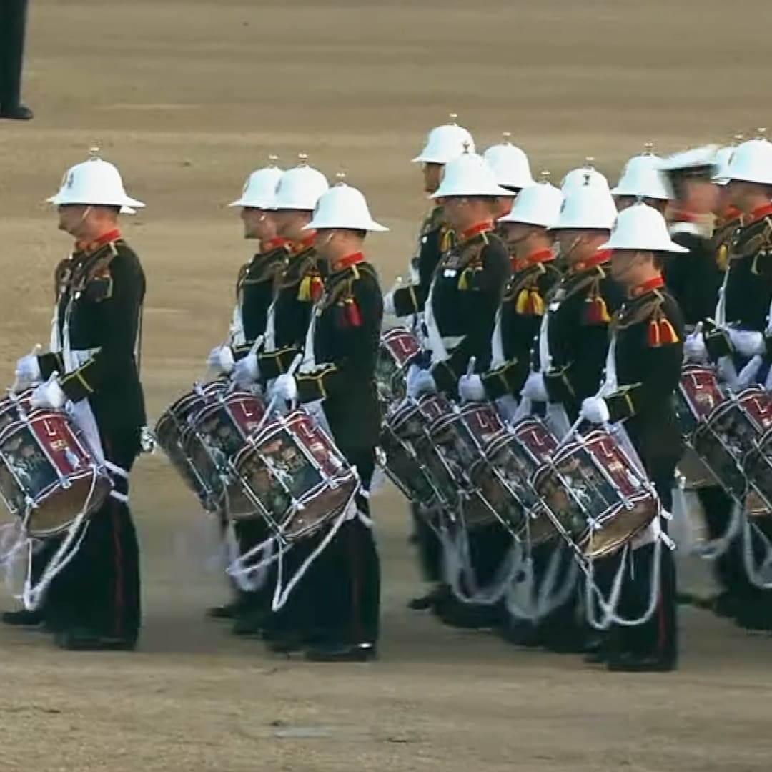 A military drum line performing at an outdoor ceremony or parade on sandy ground with young adult male soldiers in dark green uniforms and white pith helmets.