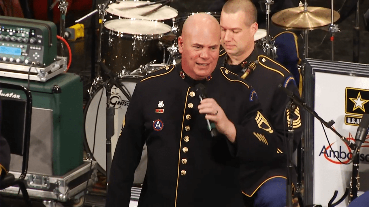 Adult male performers in formal military uniforms with gold buttons and insignia, standing on stage with a drum set behind them.