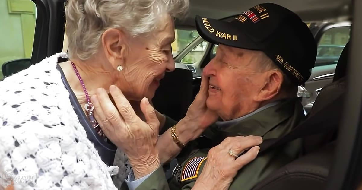 An elderly couple in a car, possibly at home; woman wears white lace with bead necklace, man is veteran wearing military-style jacket with patches and cap.