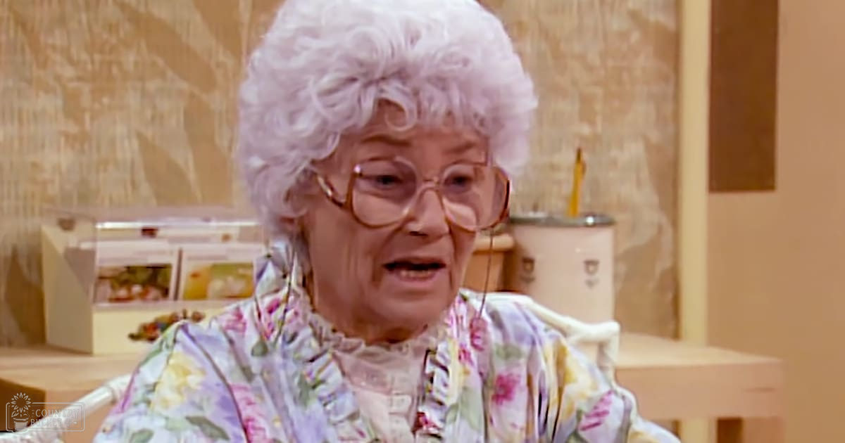 An older adult female in a domestic setting wearing a floral print blouse with long sleeves and large-rimmed glasses; wicker chair and light-colored wallpaper suggest a home environment.