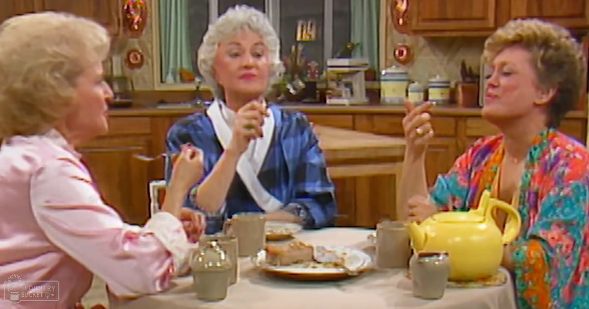 An older adult female in a pink ruffled garment with an older adult female in a blue plaid dress and another wearing a bright patterned robe, gathered around a wooden kitchen counter with jars, plants, and remnants of food suggesting a cozy homey environment.