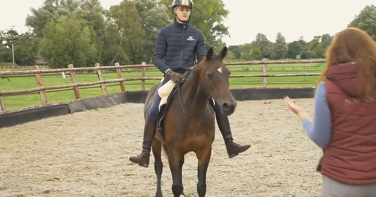 A young adult male rider in a dark blue puffer jacket and black pants rides a dark brown thoroughbred horse with white markings, observed by an adult female wearing a red vest over a light blue shirt in a sunny outdoor equestrian arena surrounded by trees.