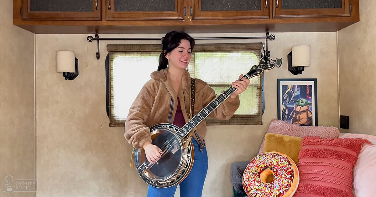 A young adult female plays a banjo inside an RV with wooden cabinets, wall-mounted sconces, and pink pillows in the foreground.