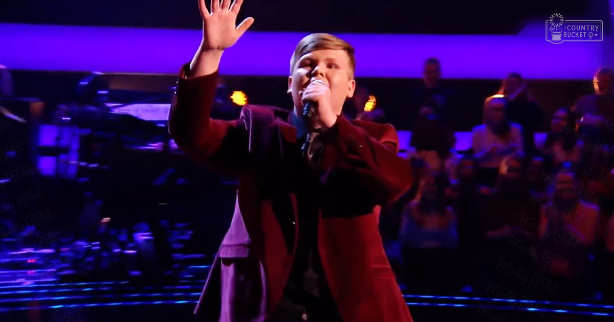 A young adult male performer wearing a dark red overcoat and white shirt is on stage at what appears to be a talent show or similar event, with an audience of adults in the background illuminated by purple lighting.