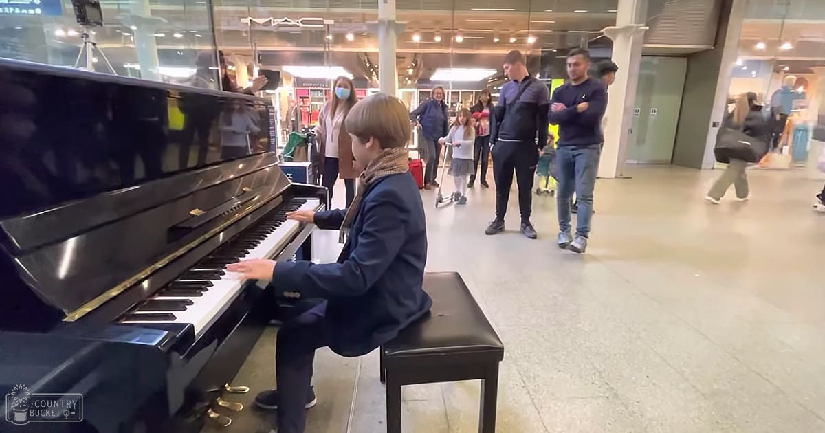 A young child playing an upright piano in a well-lit indoor public space resembling a shopping mall, surrounded by adults and other children.