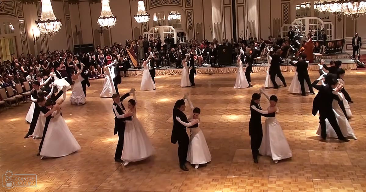 A ballroom dance performance in an elegant hall with male dancers in black tuxedos and female dancers in white satin ball gowns, ornate chandeliers hanging from the ceiling, and a band playing violins and cellos.