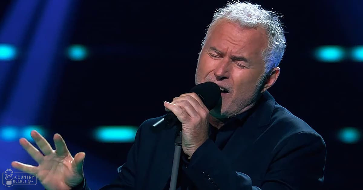 A middle-aged adult male performing on a stage with dark-colored, fitted formal attire under blue stage lighting.