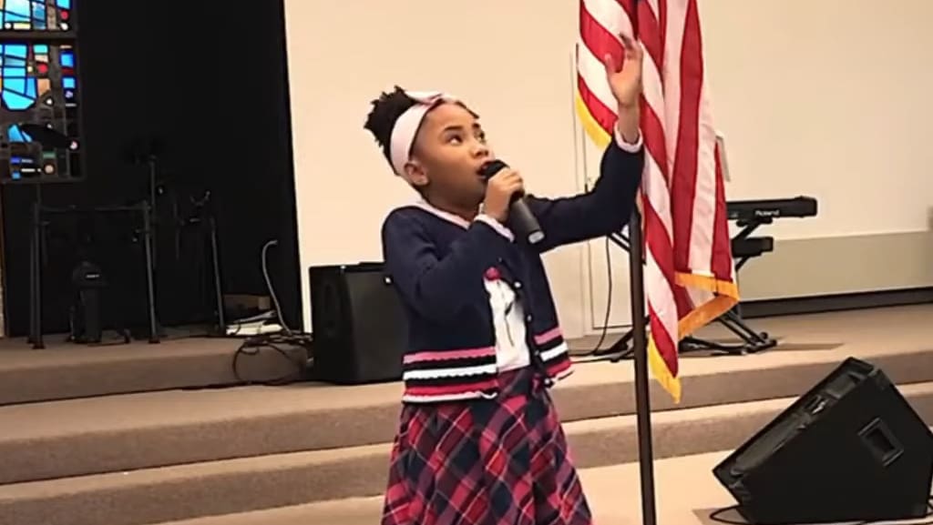 A child singing in front of an American flag at a church or community hall wearing a navy blue cardigan with white and pink trim and a plaid skirt.